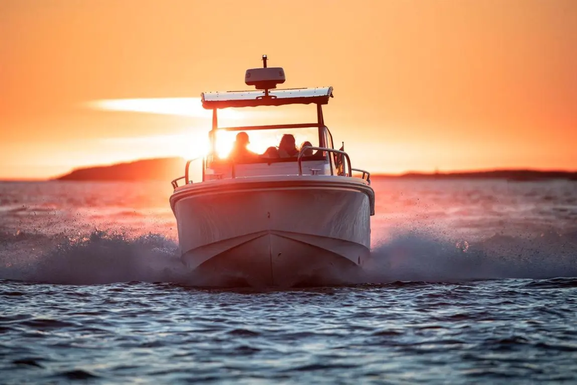 A boat cruising on water at sunset, leaving a wake, with four passengers enjoying the view against a vibrant orange sky.